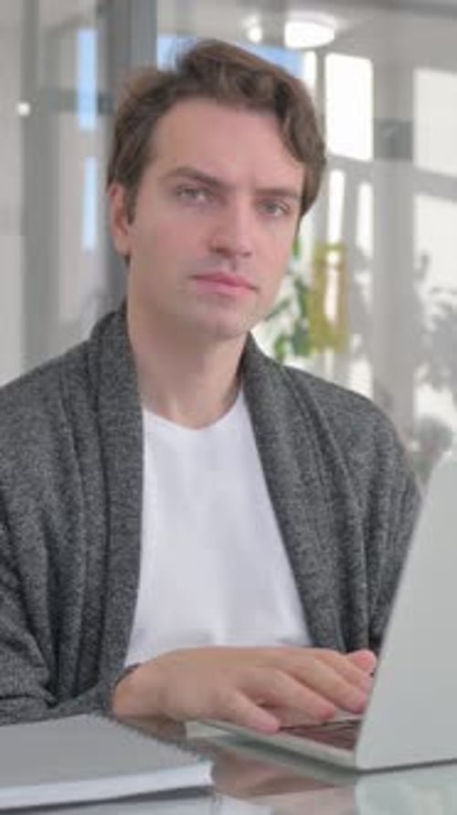 Young Man Looking at Camera while Working on Laptop in Office, vertical video