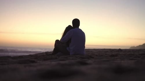 Static handheld shot of a couple in love in silhouette sitting on the beach in front of the sea with