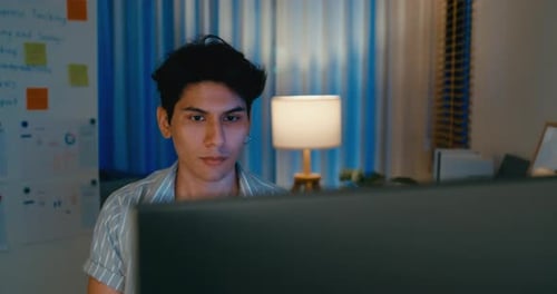 Asiatische Männer nutzen Computerfernarbeit am Schreibtisch im Wohnzimmer im Heimbüro, Überstunden am Abend.