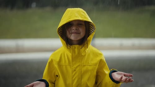 Porträt eines glücklichen kleinen Teenagers in einer gelben Jacke, das im Regen steht und fängt