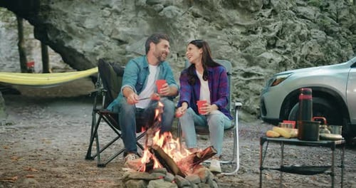 Attaractive young woman and bearded man sitting in forest near rock, roasting sausages by bonfire