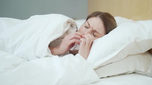 Femme blonde malade se mouchant, éternuant et toussant allongée sur le lit chaud de la chambre