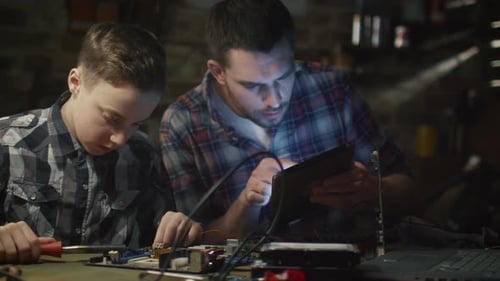 Father and son are soldering a circuit board in a garage at home.