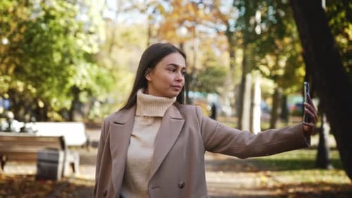 Young Beauty Woman Walks in Coat in Autumn Park Holds Phone in Hand and Takes Picture of Herself on