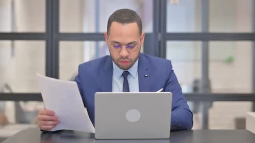 Man Working on Laptop with Paper Documents