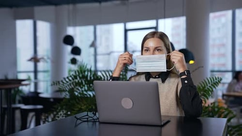Girl in office puts a mask on and fits it to her face. Long-haired female employee wears mask