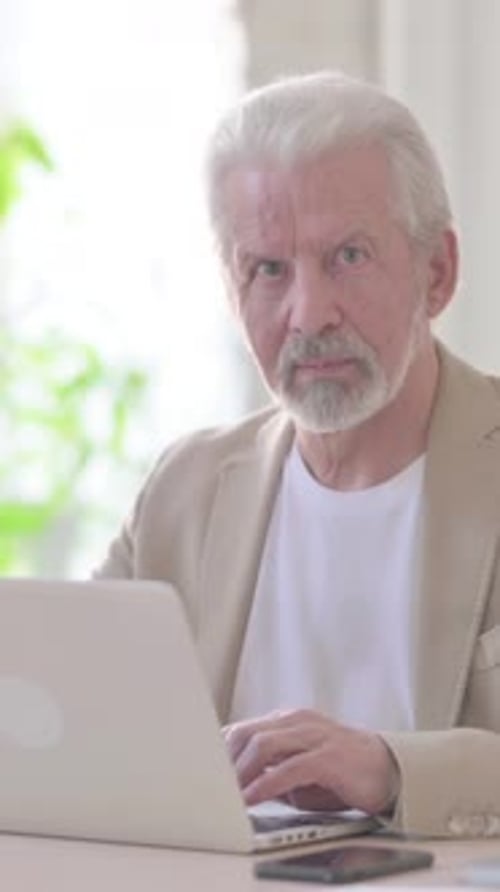 Old Man Looking at Camera while using Laptop in Office, Vertical Video