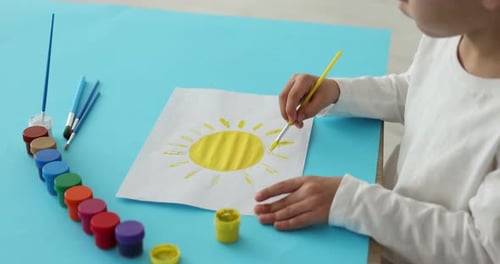 Kleiner Junge zeichnet ein Bild mit Farben und Pinsel am Tisch drinnen, Nahaufnahme. Kamera bewegt sich