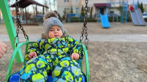 Ruhiges ernstes Kind, das auf dem Spielplatz schaukelt. Mutter klatscht dem Sohn zu, um ihn aufzumuntern, und das Baby lächelt
.