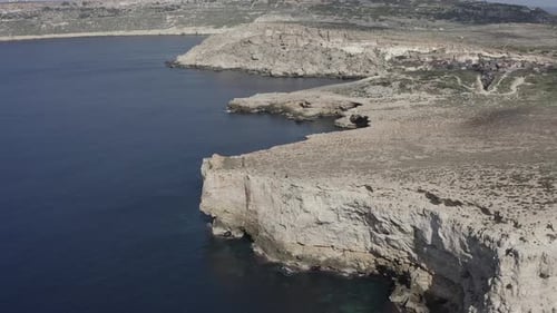 A drone flies backward along Malta’s dramatic dry cliffs, revealing rocky coasts, blue-grey sea, and