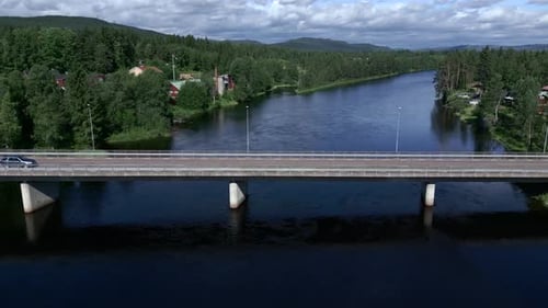 A car driving on a small bridge over a river surrounded by trees with mountains in the far backgroun