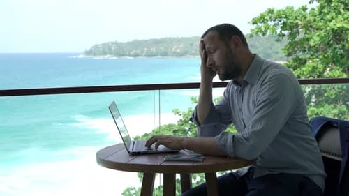 Man using laptop on balcony overlooking tropical ocean
