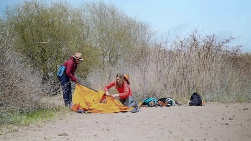 Two Women Setting Up Tent Side View Near Lake Assembling Shelter and Organizing Gear From Backpacks