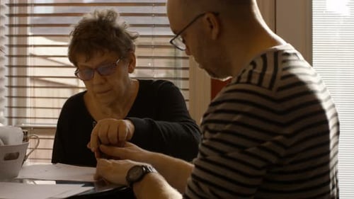 Senior Woman Reviews And Signs Document With Help From Caregiver By Kitchen Table At Home.