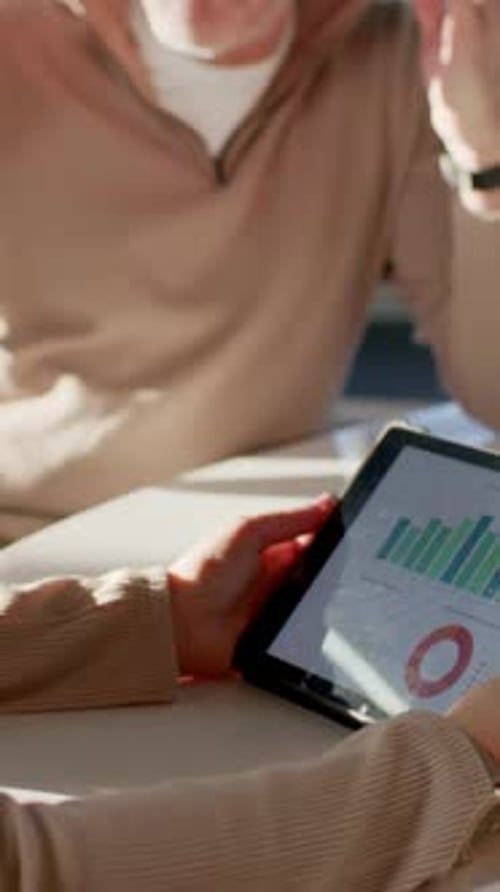 Vertical video: Leaning, man in tan top talking about bar or donut charts on tablet during meeting