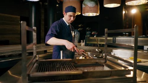 in a Japanese Restaurant Chef in a Blue Uniform Fries Fish on the Grill in the Kitchen Take on Plate