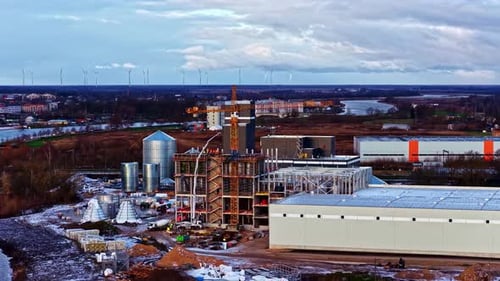 A Large Industrial Complex Rises Beside a Winding River, With Cranes, Silos, and Unfinished
