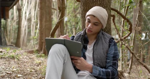 In forest, young man pondering ideas while writing in notebook under tree