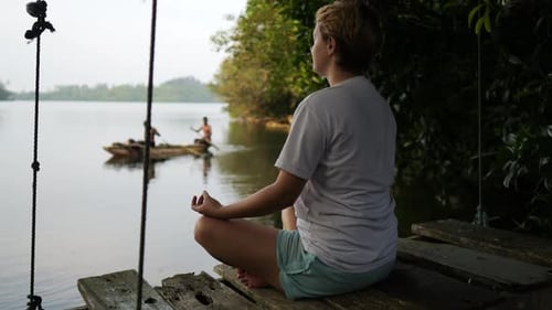 Frau meditiert, während sie auf einem hölzernen Pier mit See im Hintergrund sitzt