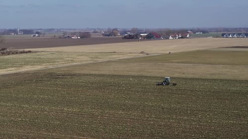 Small tractor drives over field in agriculture and a couple of farmhouses - Aerial view drone flight