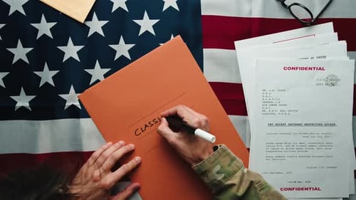 Hand Writes 'Classified' on Folder on American Flag