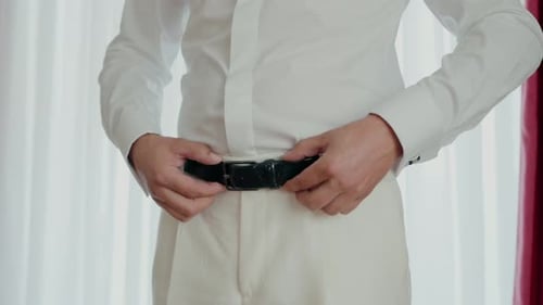 groom tightening his black belt over a white suit with a plain white shirt in preparation for his we