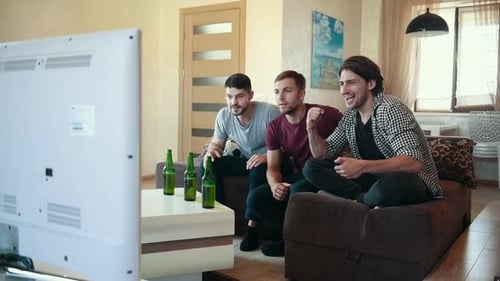 Three adult male friends with beer on table watching soccer indoors and celebrating victory