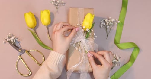 Florist decorating gift box with flowers and ribbon on pink background, top view