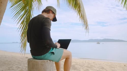 Mann sitzt mit Laptop unter einer Palme am tropischen Sandstrand und arbeitet neben der Ocean Horizon-Fernbedienung