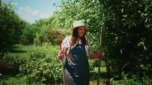 Femme jardinière portant un chapeau de paille et un tablier tenant des outils dans un jardin verdoyant