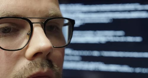 A man with glasses in close-up, focused, with a large display behind him showing AI programming code