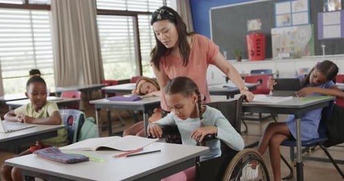 Diverse female teacher teaching schoolgirl in wheelchair reading in classroom at elementary school