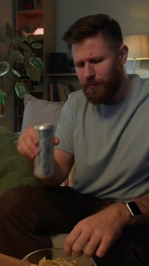 Vertical of Young Man Eating Potato Chips and Drinking Soda at Home in Evening