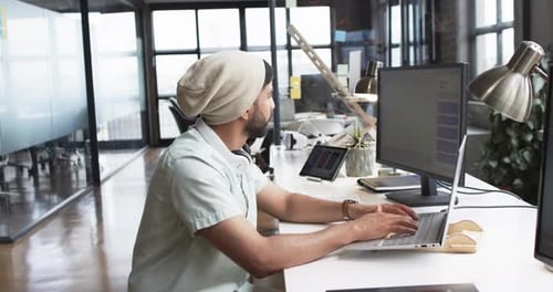 Young Asian man works on laptop in a modern business office setting, wearing headphones and a beanie