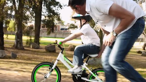 Father teaches son to ride bike on park pathway with safety helmet steadicam shot