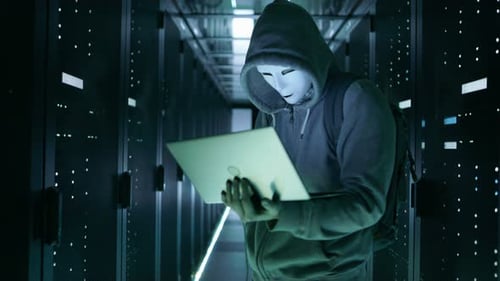 Close-up Shot of a Masked Hacker in a Hoodie Standing in the Middle of Data Center full of Rack Ser