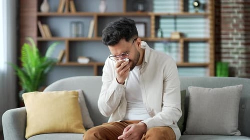 Young handsome bearded man with an allergy sneezes sitting on sofa in living room at home. Sick