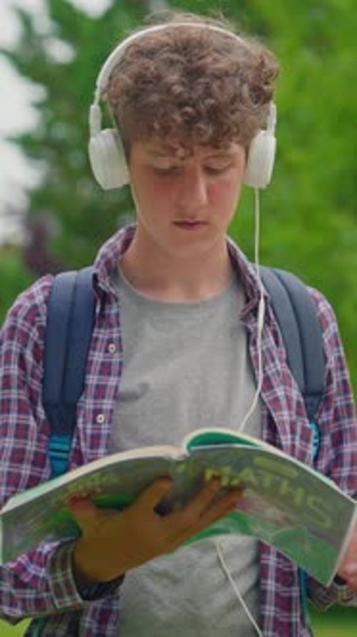Vertical Video Curly Teenage Boy Listening to Music on Headphones and Reading a Book While Sitting