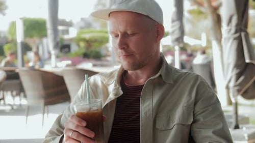 A Young Man is Sitting in a Cafe in the Fresh Air and Drinking Iced Coffee