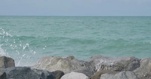Ocean waves crash against large coastal rocks, sending white spray into the air on a bright day