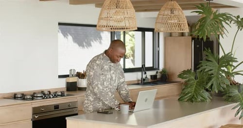 Military man using laptop and smartphone in modern kitchen, working remotely