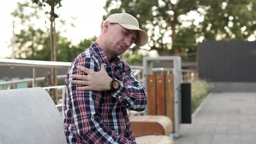 Man Rubbing Sore Shoulder Muscle on Park Bench