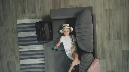 Child Awaits Vacation Laying on Couch with Suitcase