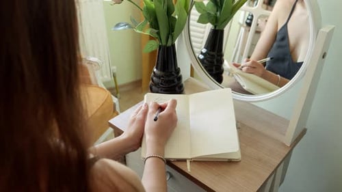 Closeup of young woman sitting at mirror in bedroom and writing in notebook