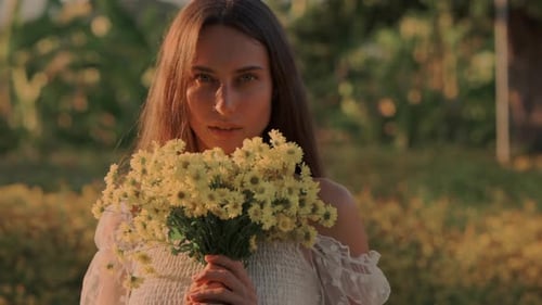 A Woman Standing in a Sunlit Field Gracefully Holding a Beautiful Yellow Flower Bouquet