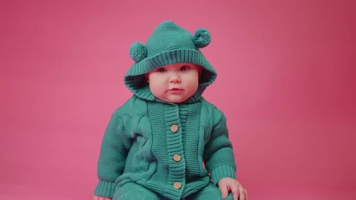 Portrait of a Cute Baby in a Warm Knitted Jumpsuit with a Hood on a Pink Background