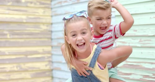Portrait of happy caucasian sister carrying her brother with sunglasses over beach house