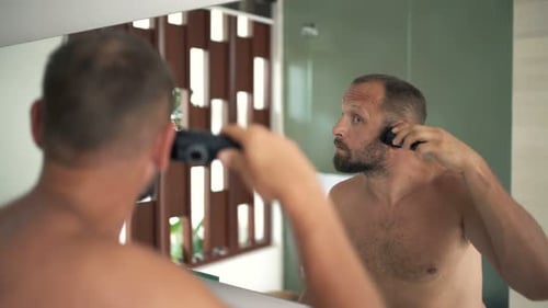 Young man shaves his beard with an electric razor in the bathroom mirror