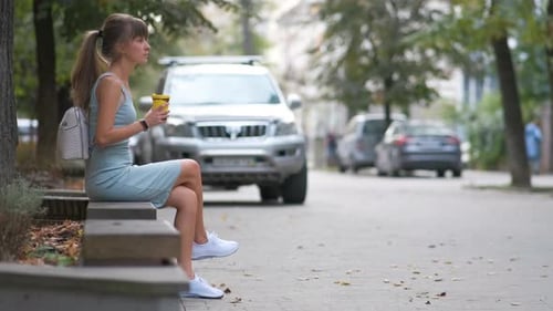 Young Woman Drinking Coffee From Paper Cup Sitting on City Street Bench in Summer Park