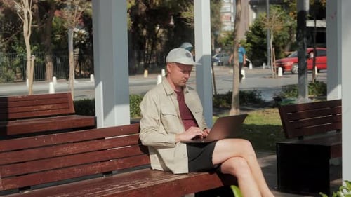 A Young Man is Sitting on a Bench in the Park and Working on a Laptop